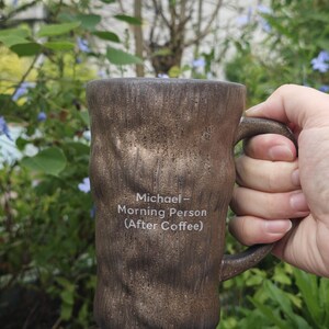 Puede incluir: Una taza de cer&aacute;mica marr&oacute;n con una superficie texturizada similar a la corteza y un asa. La parte frontal de la taza presenta un texto impreso en blanco que dice: Michael - Morning Person (After Coffee). La taza tiene una forma cil&iacute;ndrica.