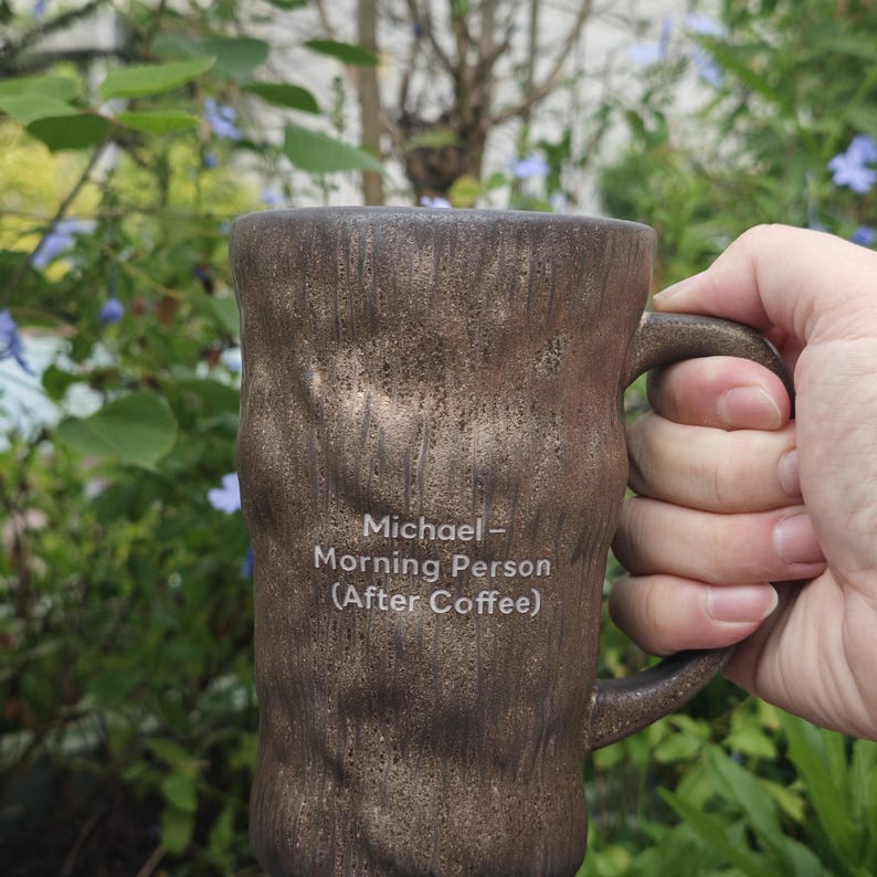 Puede incluir: Una taza de cer&aacute;mica marr&oacute;n con una superficie texturizada similar a la corteza y un asa. La parte frontal de la taza presenta un texto impreso en blanco que dice: Michael - Morning Person (After Coffee). La taza tiene una forma cil&iacute;ndrica.
