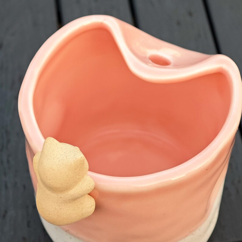 May include: A pink ceramic planter with a light beige squirrel-shaped accent. The planter has a heart-shaped top and a small hole. The bottom of the planter is white with speckled details. The planter is sitting on a dark surface.