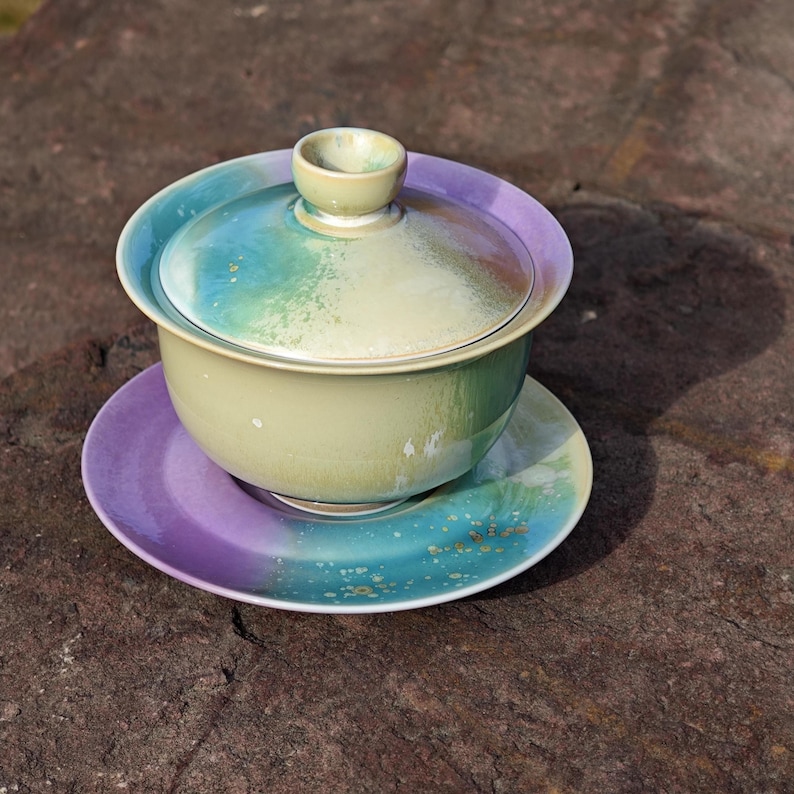May include: A ceramic tea set with a lid, bowl, and saucer. The set features a gradient of colors, including green, blue, and purple. The tea set is designed with a lid and a small handle on top. The saucer is round and has a matching color scheme.