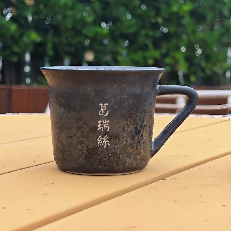 May include: A dark brown ceramic mug with a handle, featuring white Chinese characters. The mug has a textured surface and a slightly flared rim. It sits on a light yellow wooden surface, with a blurred green background.