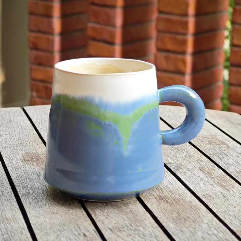 May include: A ceramic mug with a blue, green, and white color scheme. The mug has a rounded handle and a wide opening. The mug is sitting on a wooden table. The mug is approximately 4 inches tall.