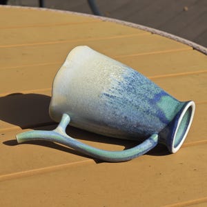 May include: A ceramic mug with a unique branch-shaped handle. The mug features a gradient of colors, transitioning from white to blue and green. The mug is resting on a yellow-brown surface.