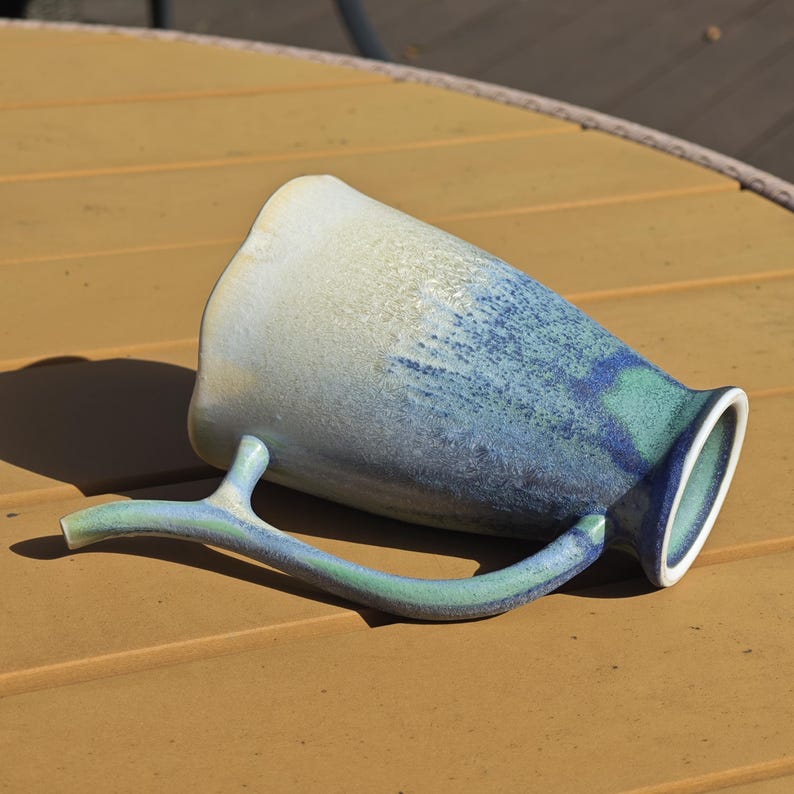 May include: A ceramic mug with a unique branch-shaped handle. The mug features a gradient of colors, transitioning from white to blue and green. The mug is resting on a yellow-brown surface.