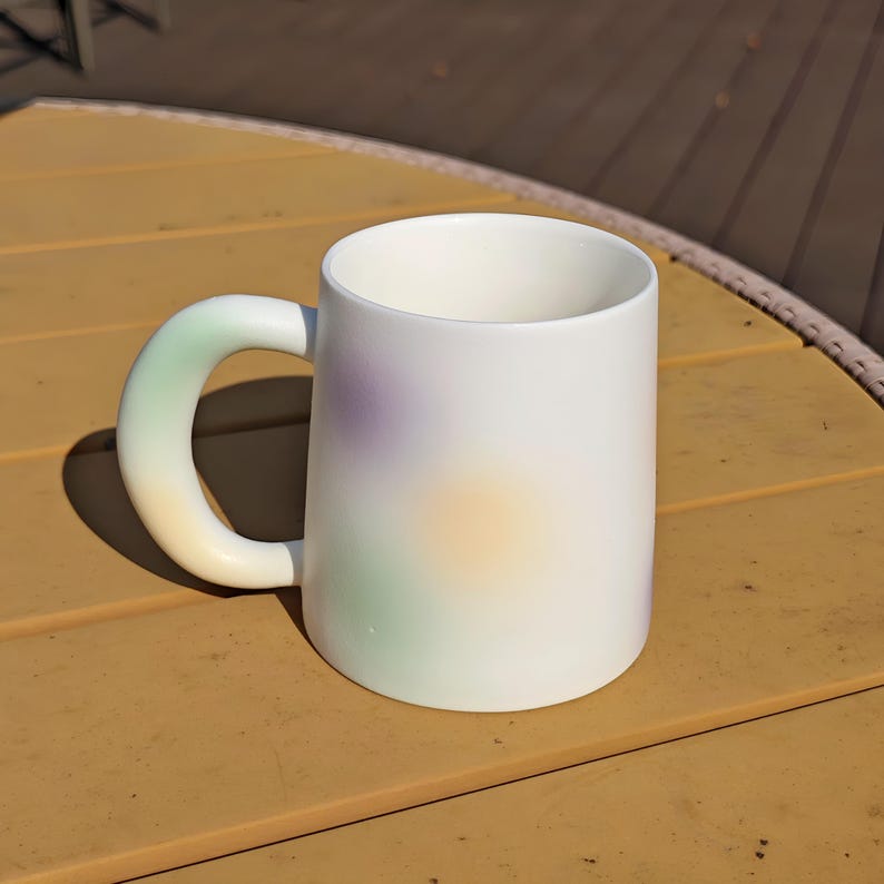 K&ouml;nnte beinhalten: Wei&szlig;er Keramikbecher mit rundem Griff. Der Becher hat einen weichen Farbverlauf aus Gr&uuml;n, Gelb und Lila. Der Becher steht auf einer gelben Oberfl&auml;che.