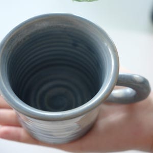 May include: A blue ceramic mug with a rounded handle. The interior has concentric circular ridges, and the exterior has horizontal ridges. The mug is handmade. The mug is held in a person's hand, showing its size and shape.