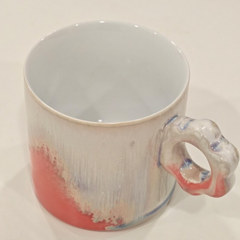 May include: A ceramic mug with a unique handle design. The mug features a white interior and a light gray exterior with red and blue accents. The handle is a sculpted, flower-like shape. The mug is cylindrical and appears to be handmade.