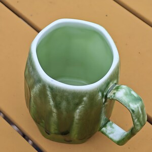 May include: A green ceramic mug with a unique, textured exterior and a white rim. The mug has a rounded, irregular shape and a sturdy handle. The interior is a solid, lighter shade of green. The mug is sitting on a yellow surface.