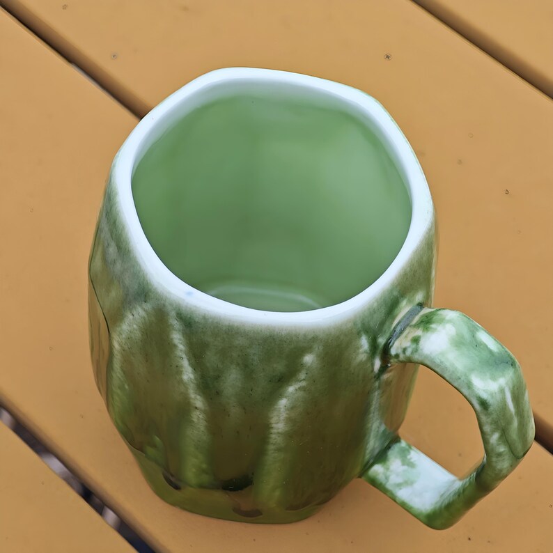May include: A green ceramic mug with a unique, textured exterior and a white rim. The mug has a rounded, irregular shape and a sturdy handle. The interior is a solid, lighter shade of green. The mug is sitting on a yellow surface.