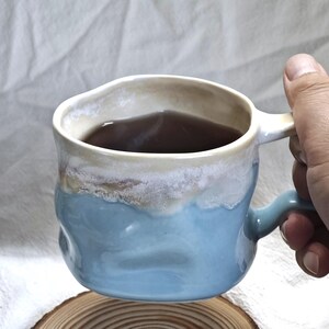 May include: A handmade ceramic mug with a unique, textured design. The mug features a light blue base, transitioning to a creamy white rim. The interior holds a dark liquid, likely tea or coffee. The mug is held by a hand, showcasing its handle.