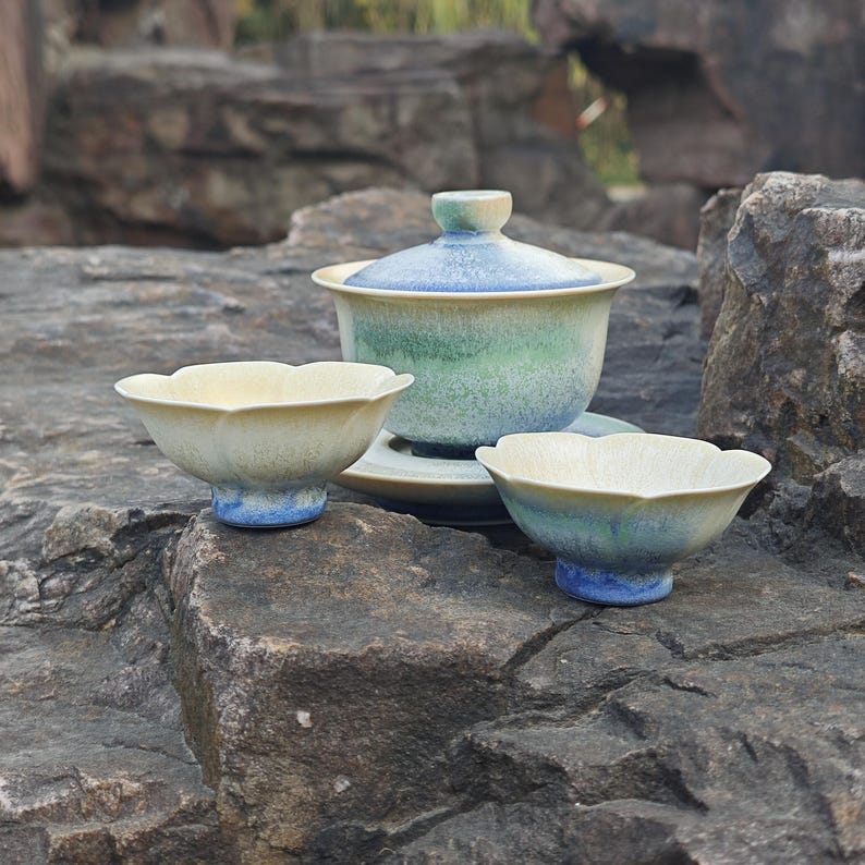 May include: A ceramic tea set with a lidded teapot, a saucer, and two small bowls. The set features a color gradient of blue, green, and cream. The bowls have a scalloped edge and sit on a blue base. The set is displayed on a rocky surface.