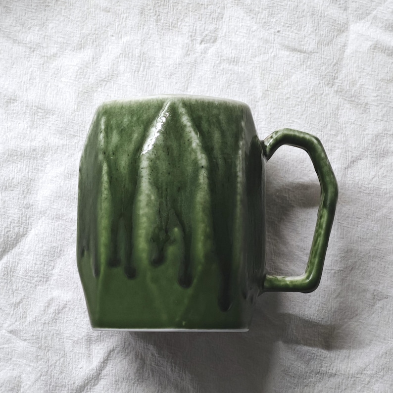May include: A green ceramic mug with a geometric design and a matching handle. The mug has a textured surface with darker green drips, creating a unique, handcrafted appearance. The mug is designed for hot beverages.