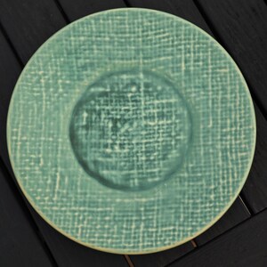 May include: A round, teal-colored plate with a textured, woven pattern. The center of the plate is a slightly darker shade of teal. The plate has a raised rim and a matte finish, suitable for serving food.