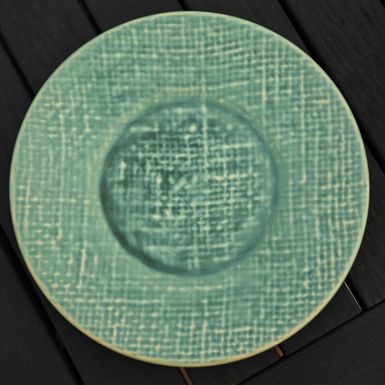 May include: A round, teal-colored plate with a textured, woven pattern. The center of the plate is a slightly darker shade of teal. The plate has a raised rim and a matte finish, suitable for serving food.