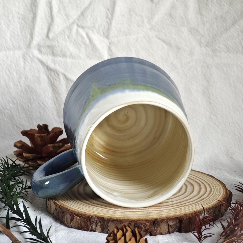 May include: A handmade ceramic mug with a blue and white glaze. The interior of the mug has a natural wood grain pattern. The mug is resting on a wooden coaster. The handle is blue.