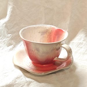May include: A ceramic teacup and saucer set. The cup and saucer are a blend of white, pink, and red hues. The cup has a curved handle and sits on a matching saucer. The set is likely handmade and suitable for serving tea or coffee.