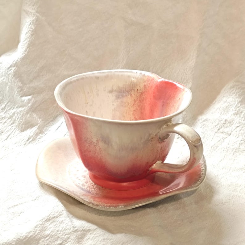 May include: A ceramic teacup and saucer set. The cup and saucer are a blend of white, pink, and red hues. The cup has a curved handle and sits on a matching saucer. The set is likely handmade and suitable for serving tea or coffee.