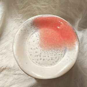 May include: A small, round ceramic plate with a white rim and a speckled, off-white interior. A splash of coral-colored glaze covers a portion of the plate's surface, creating a unique design. The plate is likely used for serving small portions of food.