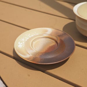 May include: A round ceramic saucer featuring a gradient glaze that transitions from a creamy off-white to a deep, earthy brown. The surface has a smooth, circular indentation in the center designed to hold a cup, with a subtle speckled texture.