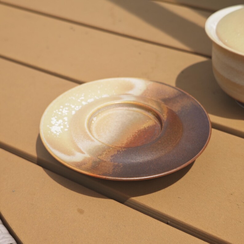 May include: A round ceramic saucer featuring a gradient glaze that transitions from a creamy off-white to a deep, earthy brown. The surface has a smooth, circular indentation in the center designed to hold a cup, with a subtle speckled texture.