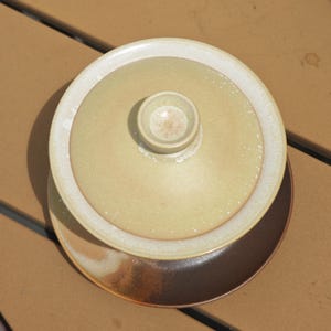 May include: A ceramic gaiwan tea set consisting of a light beige, speckled lidded bowl with a central knob, resting on a dark brown, circular saucer. The glaze on the lid and bowl features a subtle, mottled texture with lighter edges.
