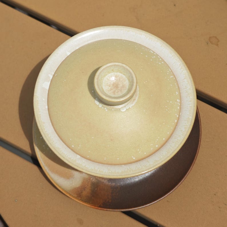 May include: A ceramic gaiwan tea set consisting of a light beige, speckled lidded bowl with a central knob, resting on a dark brown, circular saucer. The glaze on the lid and bowl features a subtle, mottled texture with lighter edges.