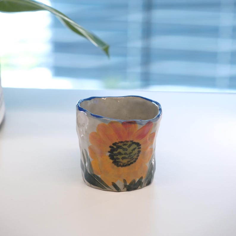 May include: A handmade ceramic cup with a rough texture and a blue rim. The cup is decorated with a vibrant sunflower design in shades of yellow, orange, and green. The cup is a unique piece of art.