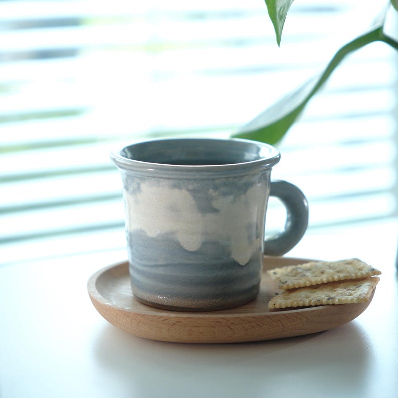 May include: A blue and white ceramic mug with a cloud design, on a small wooden plate. Two crackers are placed on the plate. The mug has a rounded handle and a flared rim. The scene is set near a window with blinds.