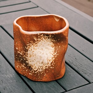 Peut inclure: Un vase en c&eacute;ramique fait &agrave; la main, de forme organique unique. L'ext&eacute;rieur est brun chaud et terreux avec un grand motif tachet&eacute; blanc cass&eacute;. L'int&eacute;rieur est brun plus fonc&eacute;. Le vase est pos&eacute; sur une surface en bois gris fonc&eacute;.