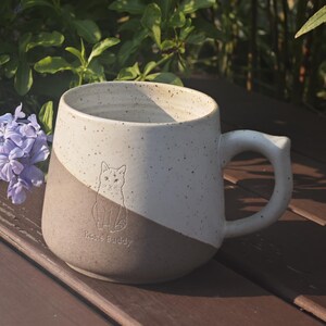 May include: A two-toned ceramic mug featuring a speckled cream upper section and a solid tan lower section. The mug displays an embossed line drawing of a cat and the text "Home Buddy" on the front, accompanied by a sturdy, curved handle.