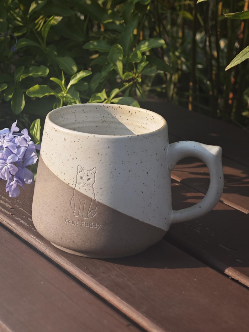 May include: A two-toned ceramic mug featuring a speckled cream upper section and a solid tan lower section. The mug displays an embossed line drawing of a cat and the text "Home Buddy" on the front, accompanied by a sturdy, curved handle.