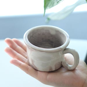 May include: A small, handmade ceramic mug with a rounded body and a sturdy handle. The mug has a light beige glaze with subtle brown marbling. The interior shows a textured pattern. The mug is held in a hand, suggesting its size and use.