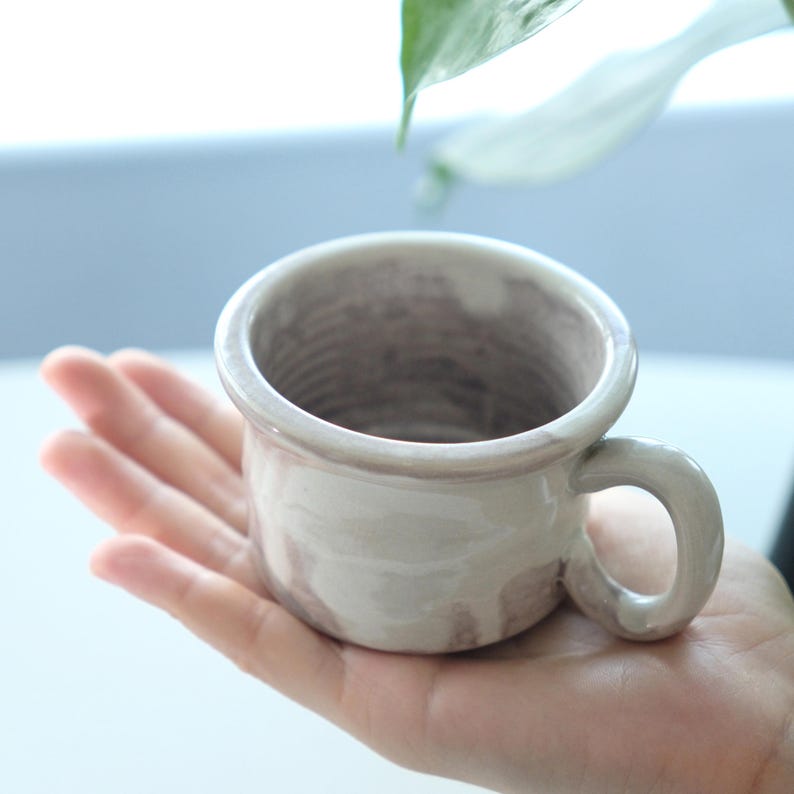 May include: A small, handmade ceramic mug with a rounded body and a sturdy handle. The mug has a light beige glaze with subtle brown marbling. The interior shows a textured pattern. The mug is held in a hand, suggesting its size and use.
