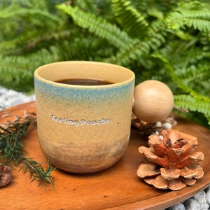 May include: A ceramic mug with a gradient design, transitioning from tan to blue, filled with dark liquid. The mug has the text "Feeling Peachy" and sits on a wooden tray with a pine cone and a wooden ball. Ferns and small rocks are in the background.