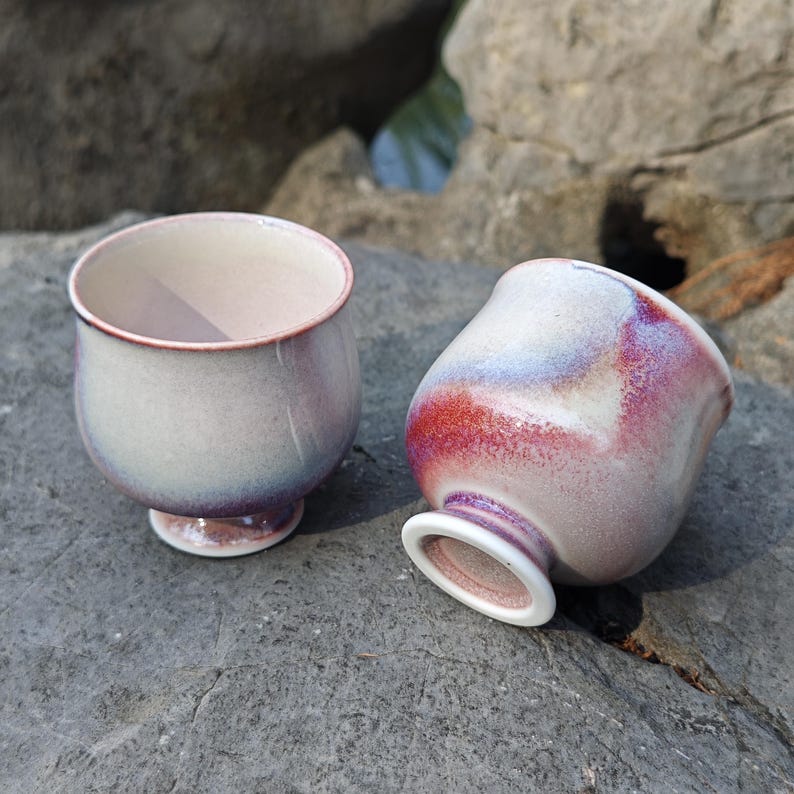 May include: Two ceramic teacups with a unique glaze. The cups have a rounded shape with a small foot and a wide opening. The glaze features a blend of colors, including white, red, and purple. One cup is upright, and the other is tilted.