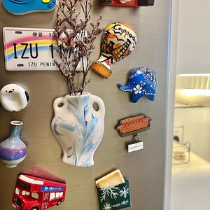 Op de afbeelding: Een verzameling decoratieve koelkastmagneten, waaronder een rode dubbeldekkerbus, een blauwe olifant met bloemmotieven en een witte vaas met blauwe wervelingen. Andere magneten hebben reisgerelateerde ontwerpen en Japanse tekst.