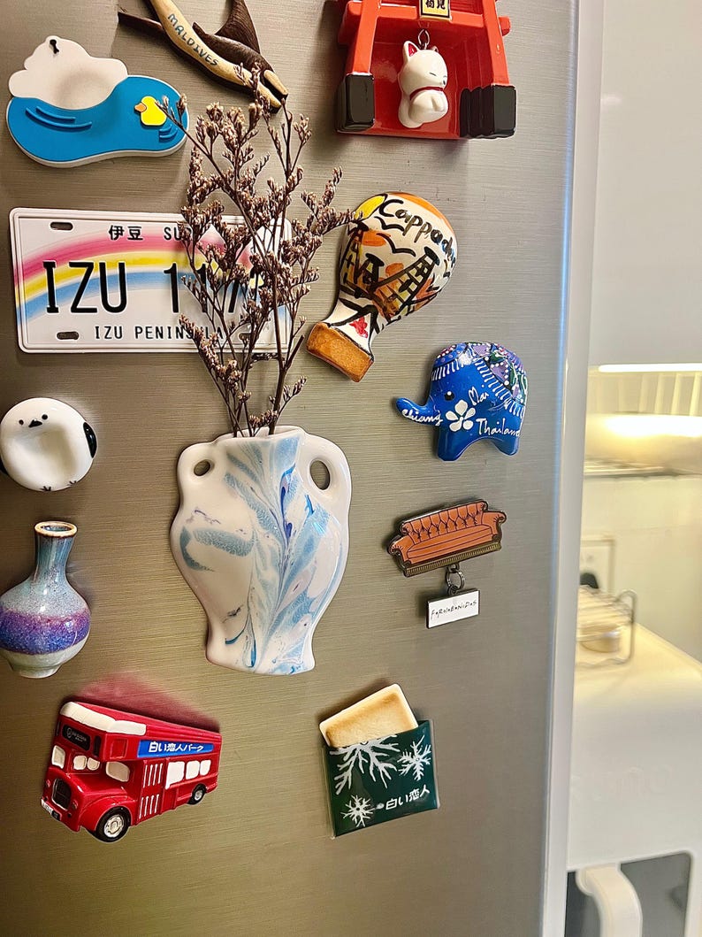 Op de afbeelding: Een verzameling decoratieve koelkastmagneten, waaronder een rode dubbeldekkerbus, een blauwe olifant met bloemmotieven en een witte vaas met blauwe wervelingen. Andere magneten hebben reisgerelateerde ontwerpen en Japanse tekst.