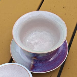 May include: A ceramic teacup and saucer set. The teacup has a wide rim and a speckled, light purple interior. The saucer is a matching color with a deep purple base. The set is on a yellow surface.