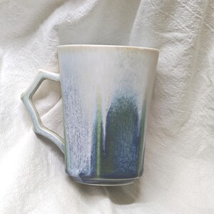 May include: A ceramic mug with a unique handle design. The mug features a gradient of colors, including white, blue, and green, creating a watercolor effect. The mug is set against a white, textured background.