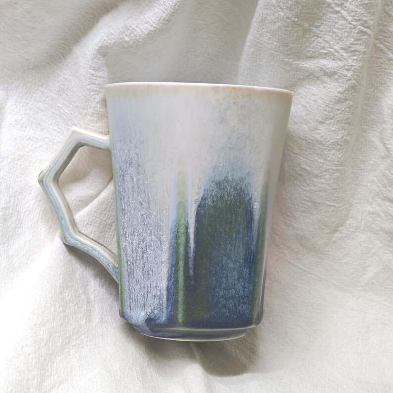 May include: A ceramic mug with a unique handle design. The mug features a gradient of colors, including white, blue, and green, creating a watercolor effect. The mug is set against a white, textured background.