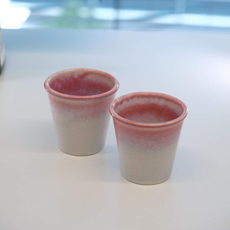 May include: Two small ceramic cups with a gradient design. The top of the cups is a reddish-pink color that fades into a creamy white. The cups are cylindrical with a slightly flared rim.