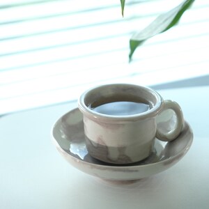 May include: A ceramic teacup and saucer set. The cup is filled with a dark liquid, likely tea or coffee. The set has a marbled, neutral color scheme with a rounded handle on the cup. The saucer is a matching, shallow dish.