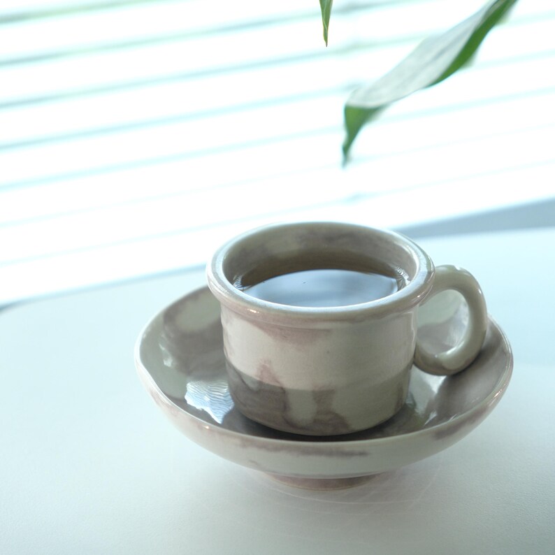 May include: A ceramic teacup and saucer set. The cup is filled with a dark liquid, likely tea or coffee. The set has a marbled, neutral color scheme with a rounded handle on the cup. The saucer is a matching, shallow dish.
