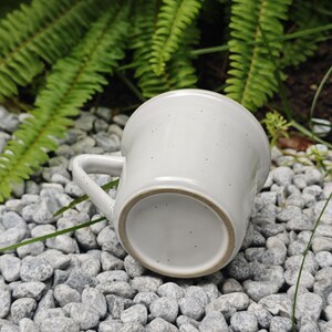 May include: A white ceramic mug with a handle, set on grey pebbles. The mug has a speckled texture and a simple, rounded design. Green foliage is visible in the background.