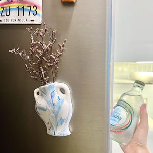 Op de afbeelding: Een kleine, witte vaas met blauwe marmerverf, met gedroogde bloemen, is bevestigd aan een koelkast. Een Japans kenteken en een decoratieve magneet staan ook op de koelkast. Een fles water wordt uit de koelkast gehaald.