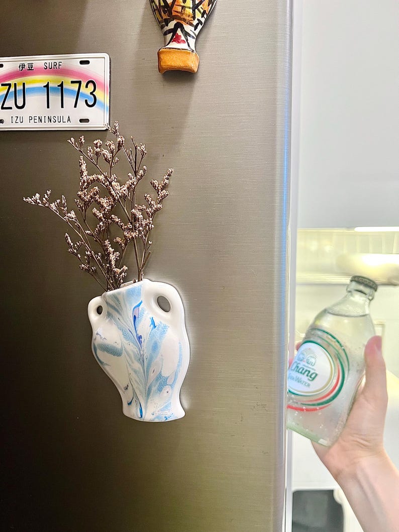 Op de afbeelding: Een kleine, witte vaas met blauwe marmerverf, met gedroogde bloemen, is bevestigd aan een koelkast. Een Japans kenteken en een decoratieve magneet staan ook op de koelkast. Een fles water wordt uit de koelkast gehaald.