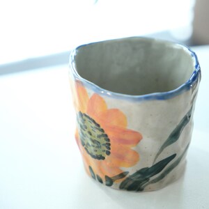 May include: A handmade ceramic mug with a sunflower design. The mug features an orange and yellow sunflower with green leaves against a cream-colored background. The rim is outlined in blue. The mug has a textured, irregular shape.