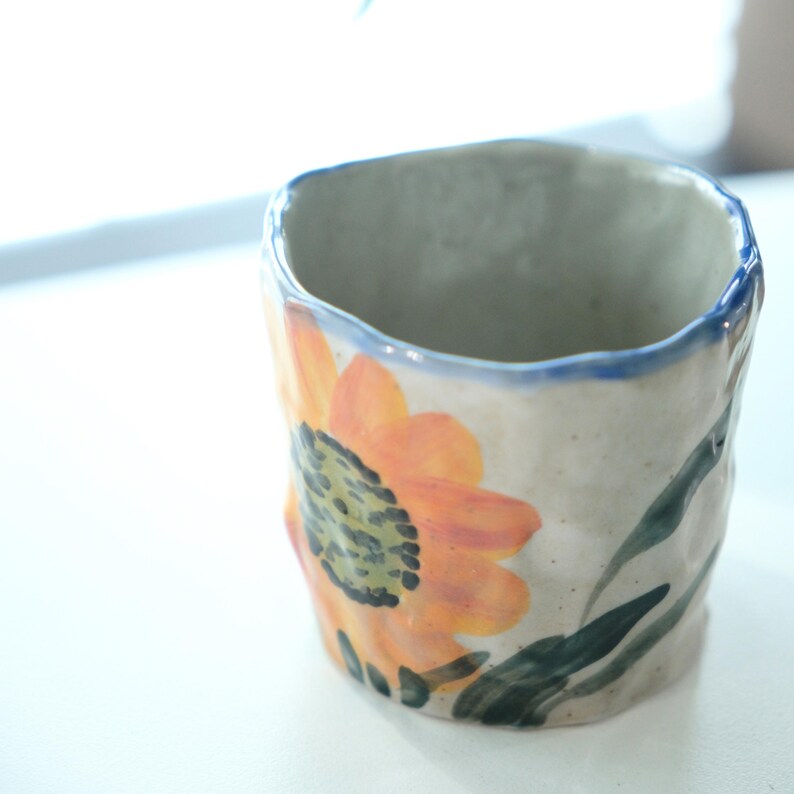 May include: A handmade ceramic mug with a sunflower design. The mug features an orange and yellow sunflower with green leaves against a cream-colored background. The rim is outlined in blue. The mug has a textured, irregular shape.