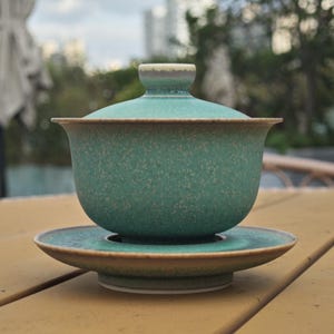K&ouml;nnte beinhalten: Ein Gaiwan-Teeservice aus Keramik, bestehend aus einer Schale, einem Deckel und einer Untertasse. Das Set hat eine gesprenkelte blaugr&uuml;ne Glasur mit matter Oberfl&auml;che. Die R&auml;nder von Schale, Deckel und Untertasse sind hellbraun abgesetzt.