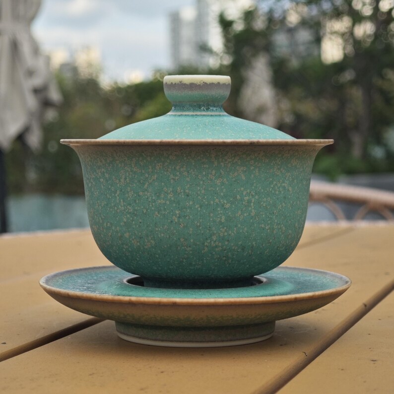 K&ouml;nnte beinhalten: Ein Gaiwan-Teeservice aus Keramik, bestehend aus einer Schale, einem Deckel und einer Untertasse. Das Set hat eine gesprenkelte blaugr&uuml;ne Glasur mit matter Oberfl&auml;che. Die R&auml;nder von Schale, Deckel und Untertasse sind hellbraun abgesetzt.